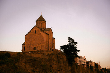Historic Metekhi church standing on a cliff above the Kura river at sunset, with traditional houses with balconies on the hillside, Tbilisi, Georgia © Hanna