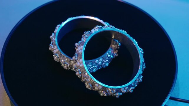 Macro close up of glowing diamond studded bangles placed on a black rotating turntable with blue neon lighting