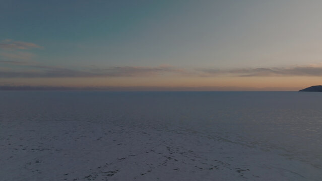 A winter aerial view across Lake Baikal near Listvyanka captures the Angara River outflow, frozen water, distant shoreline and evening colors beneath a fading sky in Irkutsk Oblast, Russia