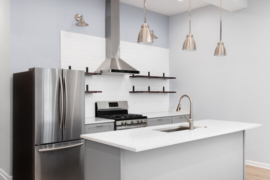 A white kitchen with grey cabinets, bronze pendant lights hanging above the island, stainless steel appliances, and white subway tile backsplash.