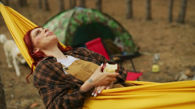 Woman relaxes in a yellow hammock at a forest campsite, holding an apple with her eyes closed while gently swaying among pine trees.