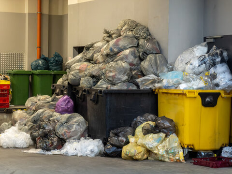 Large black and yellow industrial trash bins piled with overflowing plastic waste bags, representing urban environmental pollution, landfill management, and commercial refuse issues