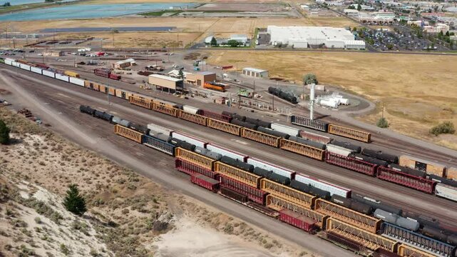 Drone flying forward over a busy freight rail yard, with long rows of boxcars, tanker cars, and auto racks on parallel tracks beside warehouses and industrial buildings, with the lake and town in the
