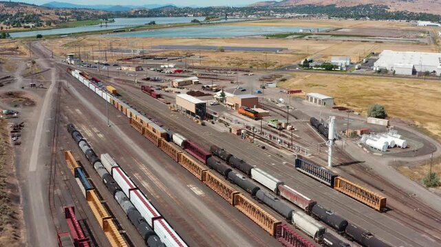 Sideways drone tracking shot over a busy freight rail yard and junction, with rows of boxcars, tanker cars, and grain hoppers lined up on parallel tracks beside warehouses, and the Klamath River and d