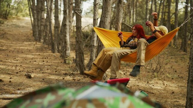 Couple chat in a hammock while camping in an autumn pine forest, eating apples as their dog walks into the campsite.