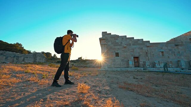 View of a photographer capturing ancient ruins at sunset. Golden light outlines the scene as the sun sets behind the historic remains.