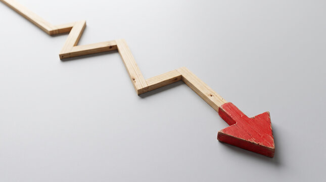 Red wooden arrow pointing down along descending line made from thin wooden strips. Falling business performance graph. Light gray background, tabletop setup. Declining sales, shrinking profits.
