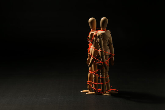 Two wooden mannequins bound together with red string on dark background. Conceptual image of toxic relationships, codependency, emotional bondage, and control.