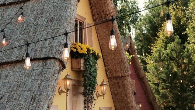 Edison bulb string lights illuminate a thatched roof cottage with yellow flowers