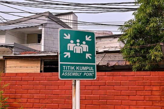 An outdoor emergency assembly point sign with white pictogram icons and Indonesian text mounted in front of a red brick wall and urban buildings.