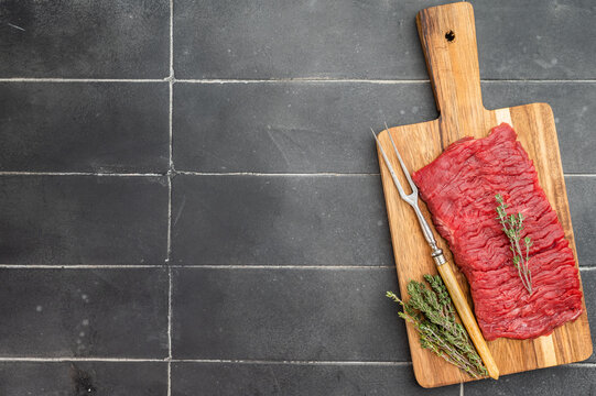 Raw beef flank steak bavette style with pronounced muscle grain on wooden board with thyme on dark tile background.
