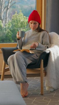 A woman reading by a large window and enjoying scenic snowy mountain view.