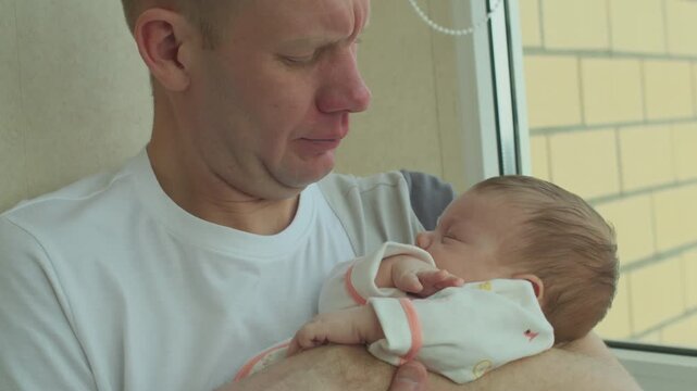 Father cradling sleeping infant daughter near window. Domestic parenting scene for fatherhood, baby care and Father Day