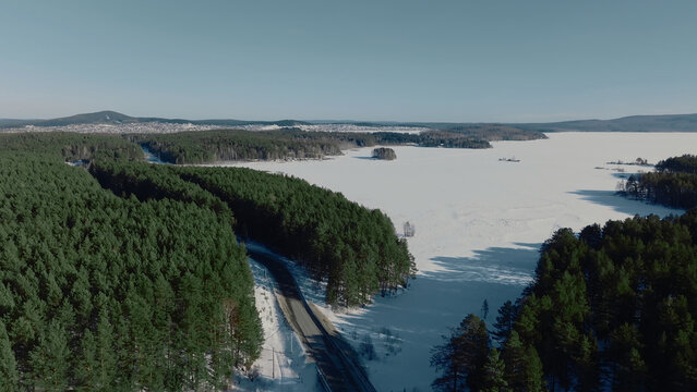 An elevated winter view follows a curved route through evergreen woodland beside an icebound lake, while snowfields and clear daylight reveal the remote landscape of the Ural Mountains, Russia