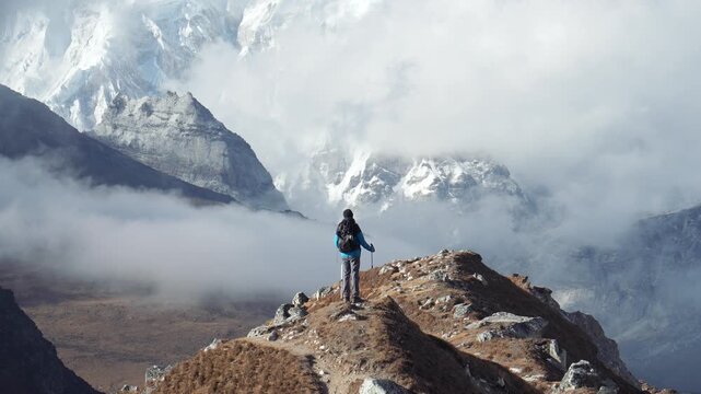 Courageous hiker with backpack and trekking poles standing on mountain ridge, looking at breathtaking panorama of snow peaks and misty clouds in beautiful himalayas of nepal