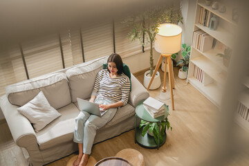 High angle view of young Asian woman typing on laptop on sofa in cozy living room with floor lamp © whyframeshot