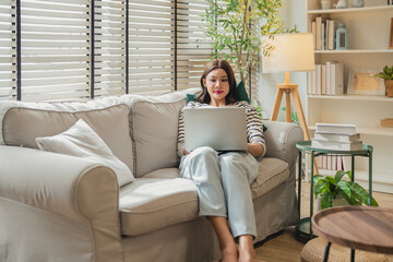 Young Asian woman working on laptop while sitting on sofa in cozy living room at home © whyframeshot