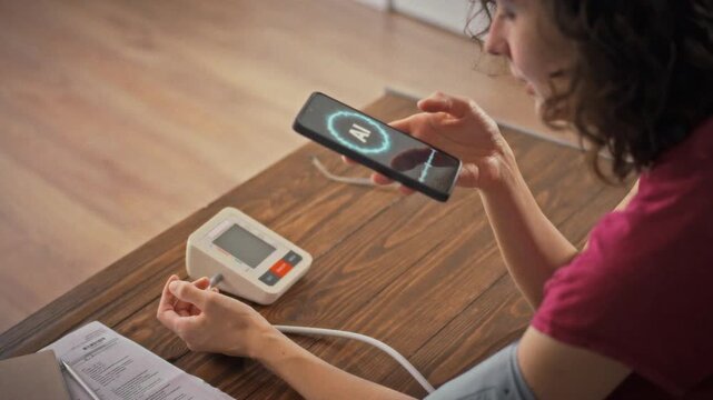 AI-powered blood pressure monitoring a young woman with a cuff on her arm talks to a voice assistant on her smartphone she is sitting next to a wooden table with health records on it