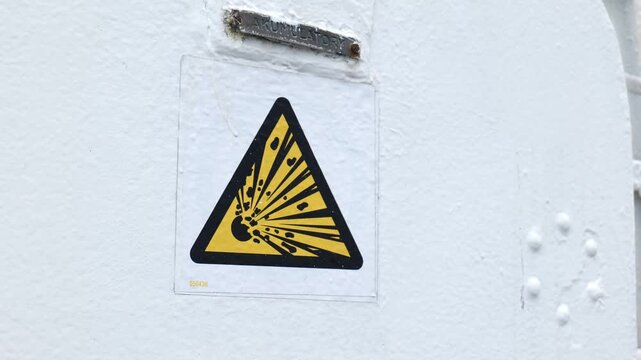 Explosion hazard warning sign, yellow triangle with black border and blast pictogram on white painted metal surface with rivets, closeup, industrial safety, danger symbol, safety signage
