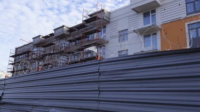 Spirals of tangled barbed wire rest atop a corrugated metal barrier in front of a modern apartment building with white and orange facades