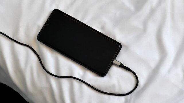 Black smartphone charging on a bed, braided charging cable plugged into the side port, screen off, lying on white crumpled bed sheets, top view closeup, overnight charging concept