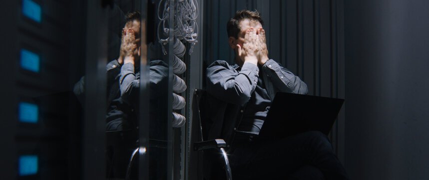 Exhausted Male Network Engineer Reacting to Critical Server Crash or Cyber Attack Late at Night. Concept of IT Burnout, Network Downtime, Data Loss Stress, and Severe Infrastructure Emergencies.