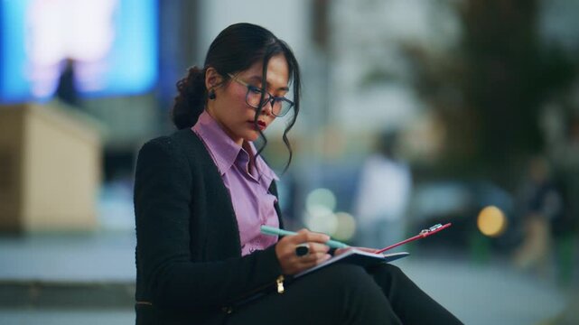 An elegant Asian businesswoman meticulously writes notes in her planner while seated outdoors. She diligently works on her tasks, focused on her document in an urban environment.