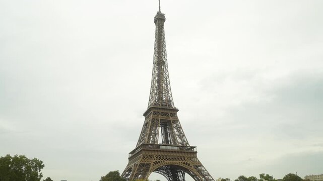 Tall iron lattice tower rising into overcast sky, filmed from ground-level wide angle showing lower arches, first observation deck, and detailed metal framework, soft diffuse daylight creates muted