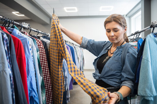 Woman inspecting yellow check shirt while browsing tidy thrift store rack, indoor retail setting rows of assorted apparel on hangers