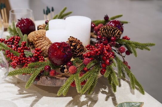 Festive table centerpiece with white candles, fir branches, red berries, pinecones and pomegranates. Christmas home decoration and holiday shopping ideas for New Year and winter celebration table sett