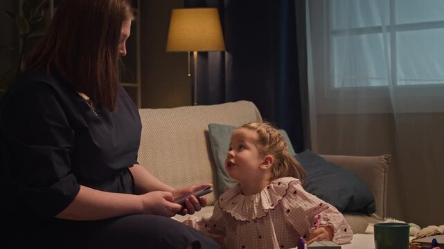 Caucasian mother setting smartphone aside to support Caucasian child daughter drawing pictures at home. Footage fits parenting apps, digital detox promos, and family lifestyle campaigns