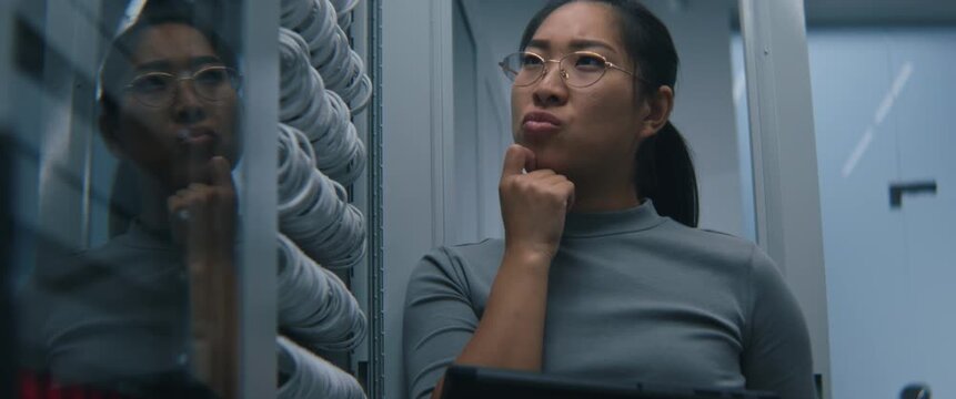 Female IT Technician Thoughtfully Analyzes Information on Tablet in Modern Server Room. Concerned Expression Conveys Infrastructure Monitoring, Technical Troubleshooting, Digital System Reliability.