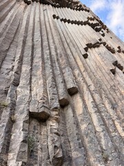 Tall gray basalt columns rise vertically across a rugged cliff face, with irregular dark sections and blue sky visible above © Anat art
