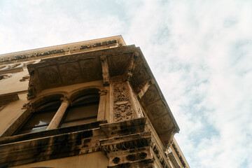 Low-angle view of ornate historic building corner with carved stone cornice and arched window under soft daylight sky, showing weathered patina on architectural details and decorative medallions, warm © Dima Anikin