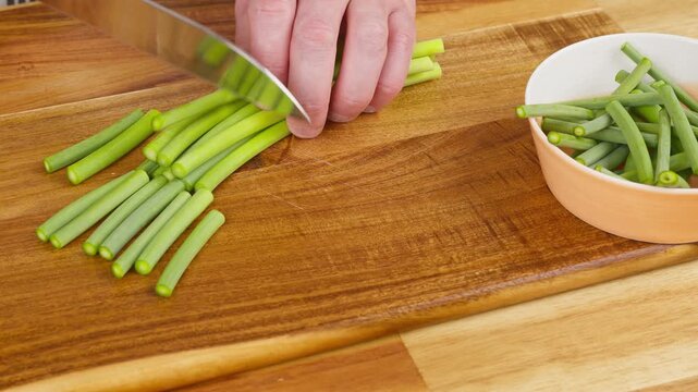 Chopping fresh green garlic sprouts into 5 cm lengths on a wooden cutting board. Real-time prep footage capturing sharp chef knife cuts for Sichuan twice-cooked pork (Suanmiao Huiguorou).