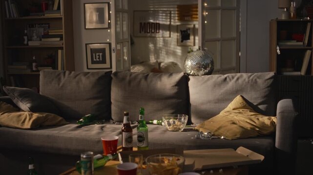 Messy living room showing scattered beer bottles, food leftovers, and disco ball after house party. Footage suitable for cleaning service or hangover remedy ad