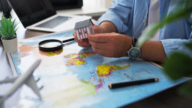 Person holding miniature house over a world map, representing immigration, international relocation, moving abroad, finding a new home, and exploring real estate opportunities in another country