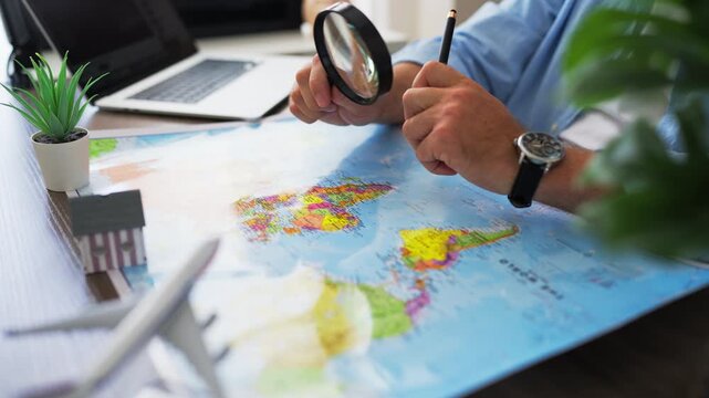 Man using magnifying glass to examine world map at desk with laptop, toy airplane, and house model. Concept of immigration, moving abroad, relocation planning, and finding new home overseas