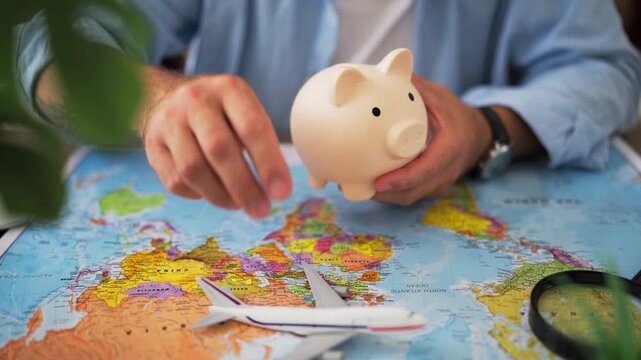 Close-up of male hands holding a piggy bank and a toy airplane over a world map, symbolizing saving money for a dream vacation, travel budget planning, and future holidays abroad