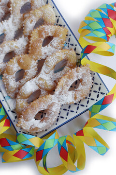 Italian sweet crisp pastry deep-fried and sprinkled with sugar in shape of Carnival mask  in a ceramic tray isolated on white background. Chiacchere or crostoli, bugie, cenci with colorful paper serpe