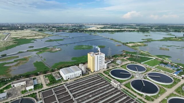 Aerial view of sewage treatment plant building with chimney releasing plume beside circular clarifier tanks and vast wetlands under blue sky, Dhaka, South Asia