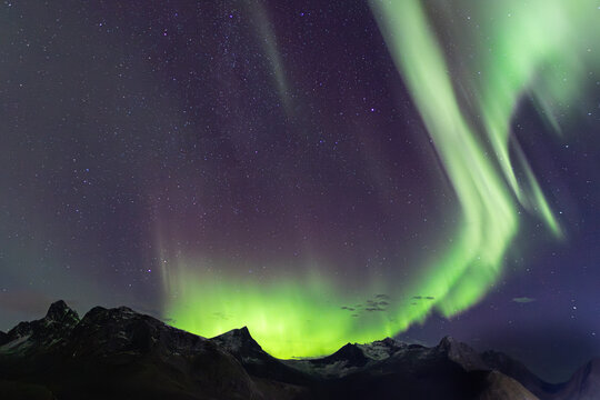 View of the Aurora Borealis illuminating the night sky above the rugged mountains of Husoy, Troms, Norway.