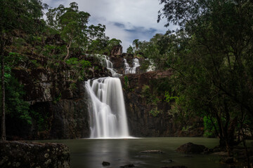 View of Cedar Creek Falls cascading over dark rocky cliffs into a tranquil pool surrounded by lush native forest in Proserpine, Queensland, Australia. © Travel 'n' Lifestyle