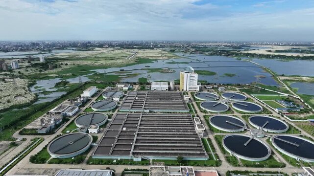 Aerial view of large sewage treatment plant with circular clarifier tanks and settling basins beside wetlands and city skyline, Dhaka, South Asia