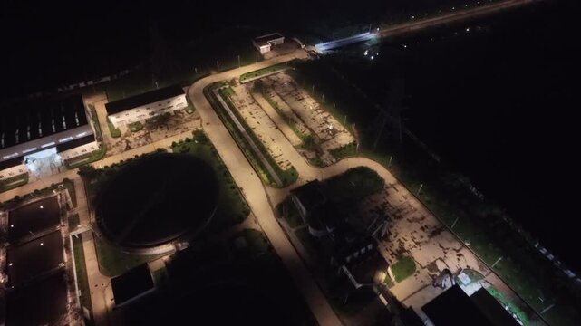 Aerial backward flight with orbit around illuminated circular clarifier tanks and buildings at night, sewage treatment plant, Dhaka, South Asia