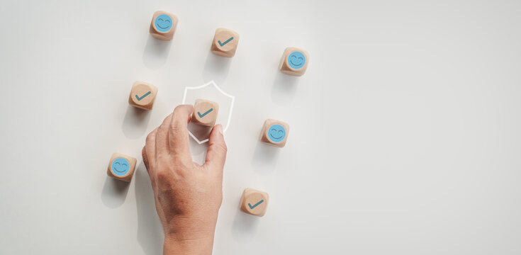 Hand placing a wooden block with check mark icon inside a protective shield among smiley face blocks on a white desk, concept of customer satisfaction, trust, and service guarantee.
