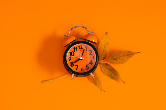 Autumn composition with orange alarm clock showing midnight and dry yellow fall leaf on vibrant orange background. Flat lay seasonal countdown concept or daylight saving time template with copy space.