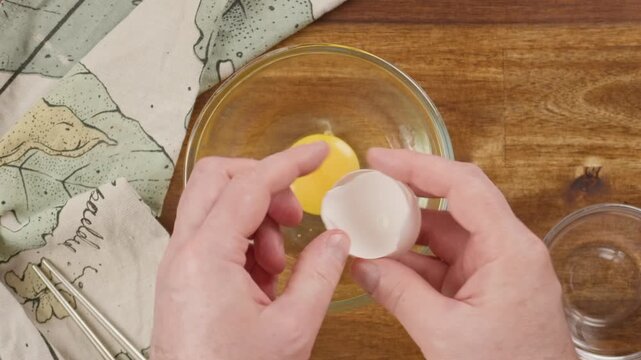 Cracking a fresh raw egg into a clear glass bowl by hand. Real-time food preparation footage capturing the shell breaking, yolk dropping, and liquid egg white flowing in a clean kitchen.