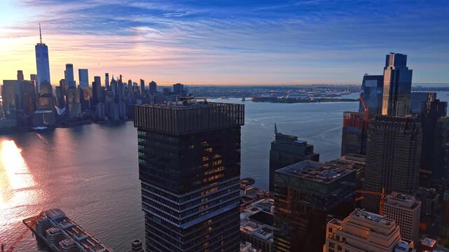 Flyover the prominent dark high-rise at the Hudson River waterfront. Iconic Manhattan skyline in the light of setting sun in the backdrop.
