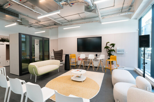 View of a modern office meeting area featuring a white sofa, yellow bar stools, a round coffee table on a patterned rug, and a large wall-mounted screen in London, United Kingdom.